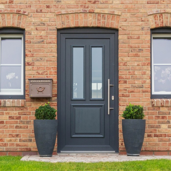 Usi-TAQ-VEKA-Softline-70- Ușă de intrare neagră modernă din PVC, cu panouri de sticlă, într-o casă din cărămidă, decorată cu două ghivece mari și o cutie poștală clasică.