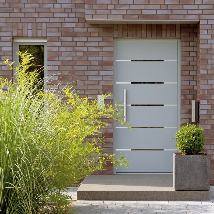 Usi-TAQ-VEKA-Softline-76-Ușă modernă de intrare gri din PVC, cu design minimalist și inserții de sticlă, montată pe o fațadă din cărămidă, înconjurată de vegetație verde.